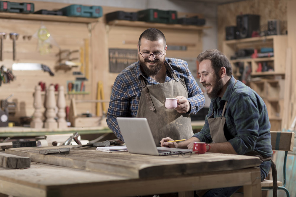 Homens olhando para um notebook e sorrido em uma pequena fábrica de móveis de madeira - contabilidade de custos